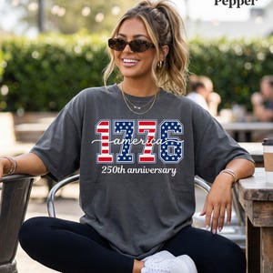 May include: A dark grey t-shirt with the text "1776 America 250th anniversary" in red, white, and blue. The design incorporates the American flag within the numbers and text. The t-shirt is a casual style.