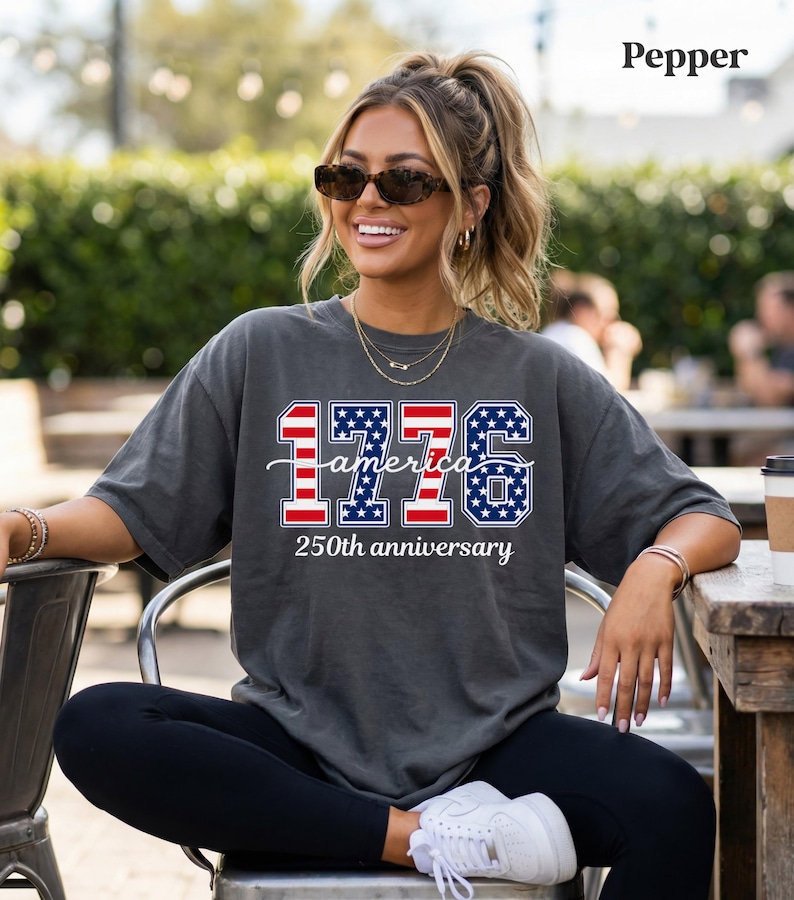 May include: A dark grey t-shirt with the text "1776 America 250th anniversary" in red, white, and blue. The design incorporates the American flag within the numbers and text. The t-shirt is a casual style.