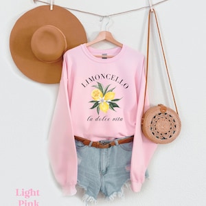 May include: A light pink sweatshirt with the text "Limoncello la dolce vita" and a graphic of lemons and leaves. The sweatshirt is hanging on a hanger with a brown hat and a brown woven bag.