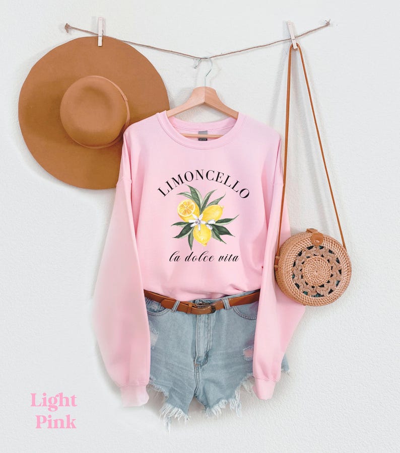 May include: A light pink sweatshirt with the text "Limoncello la dolce vita" and a graphic of lemons and leaves. The sweatshirt is hanging on a hanger with a brown hat and a brown woven bag.
