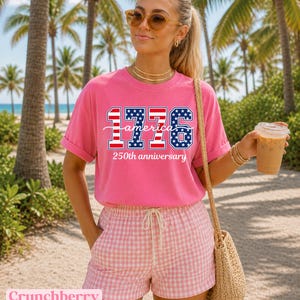 May include: A bright pink t-shirt with the text "1776 America 250th anniversary" in red, white, and blue. The person is wearing pink and white checkered shorts and sunglasses. A straw bag is over the shoulder.