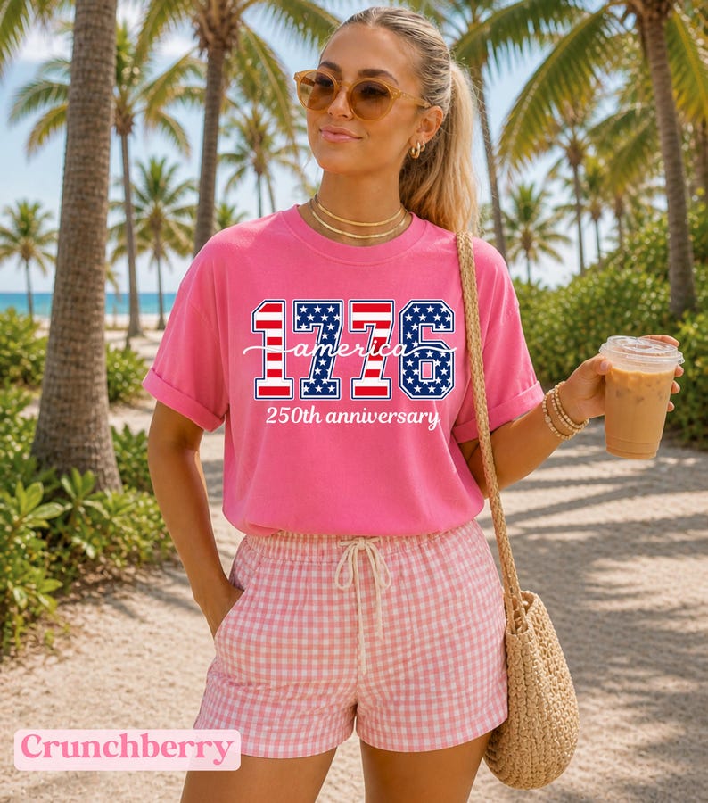 May include: A bright pink t-shirt with the text "1776 America 250th anniversary" in red, white, and blue. The person is wearing pink and white checkered shorts and sunglasses. A straw bag is over the shoulder.