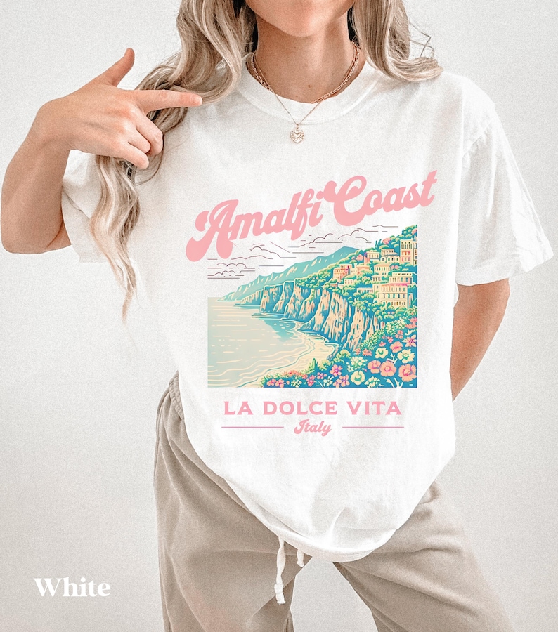 May include: White t-shirt with a graphic print of the Amalfi Coast in Italy. The graphic features a colorful illustration of the coastline with buildings, a beach, and flowers. The text "Amalfi Coast" is in pink and the text "LA DOLCE VITA Italy" is below the graphic.