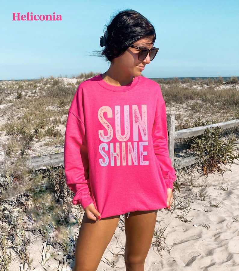 May include: A bright pink sweatshirt with the words "SUN SHINE" in a gradient font. The sweatshirt is long-sleeved and worn by a person wearing sunglasses. The word "Heliconia" is in the top left corner.