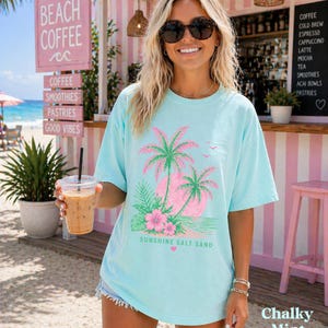 May include: A turquoise t-shirt featuring a pink and green palm tree design with the words "Sunshine Salt Sand". The image shows a beach cafe with signs advertising coffee, smoothies, and pastries. The shirt is a Chalky Mint colour.
