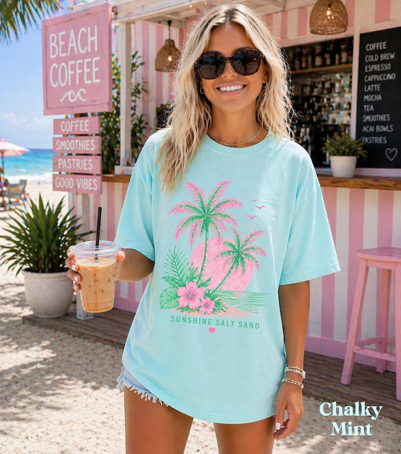 May include: A turquoise t-shirt featuring a pink and green palm tree design with the words "Sunshine Salt Sand". The image shows a beach cafe with signs advertising coffee, smoothies, and pastries. The shirt is a Chalky Mint colour.