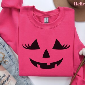 May include: A bright pink long-sleeve crewneck sweatshirt featuring a black graphic of a jack-o'-lantern face with eyelashes. The sweatshirt has a Gildan brand label at the inner neckline and is displayed on a flat surface with other items.