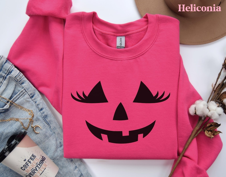May include: A bright pink long-sleeve crewneck sweatshirt featuring a black graphic of a jack-o'-lantern face with eyelashes. The sweatshirt has a Gildan brand label at the inner neckline and is displayed on a flat surface with other items.