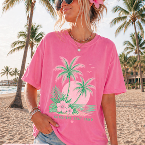 May include: Bright pink t-shirt with a tropical design. The design includes palm trees, a sunset, and the text "Sunshine Salt Sand." The shirt has a relaxed fit. The word "Crunchberry" is at the bottom of the image.