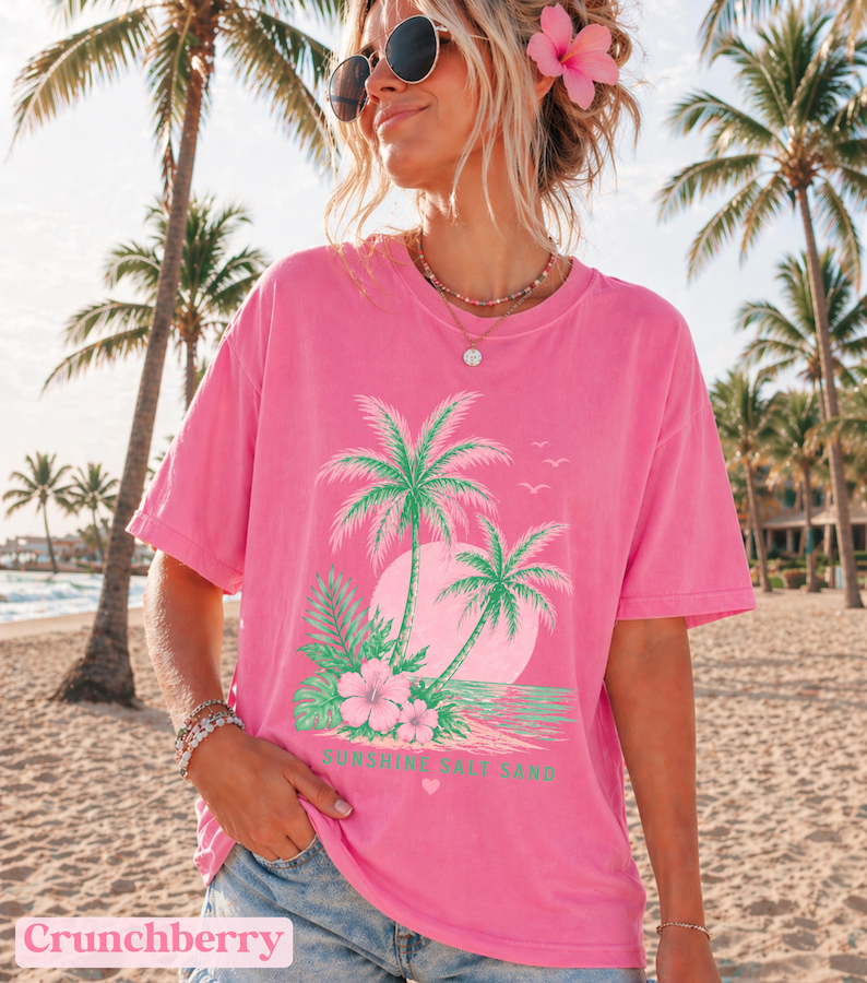 May include: Bright pink t-shirt with a tropical design. The design includes palm trees, a sunset, and the text "Sunshine Salt Sand." The shirt has a relaxed fit. The word "Crunchberry" is at the bottom of the image.