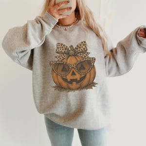 Op de afbeelding: Een lichtgrijs sweatshirt met ronde hals en een centrale afbeelding van een oranje pompoen met een zonnebril met luipaardprint en een bijpassende strik. De pompoen rust op een klein hoopje bruine herfstbladeren.