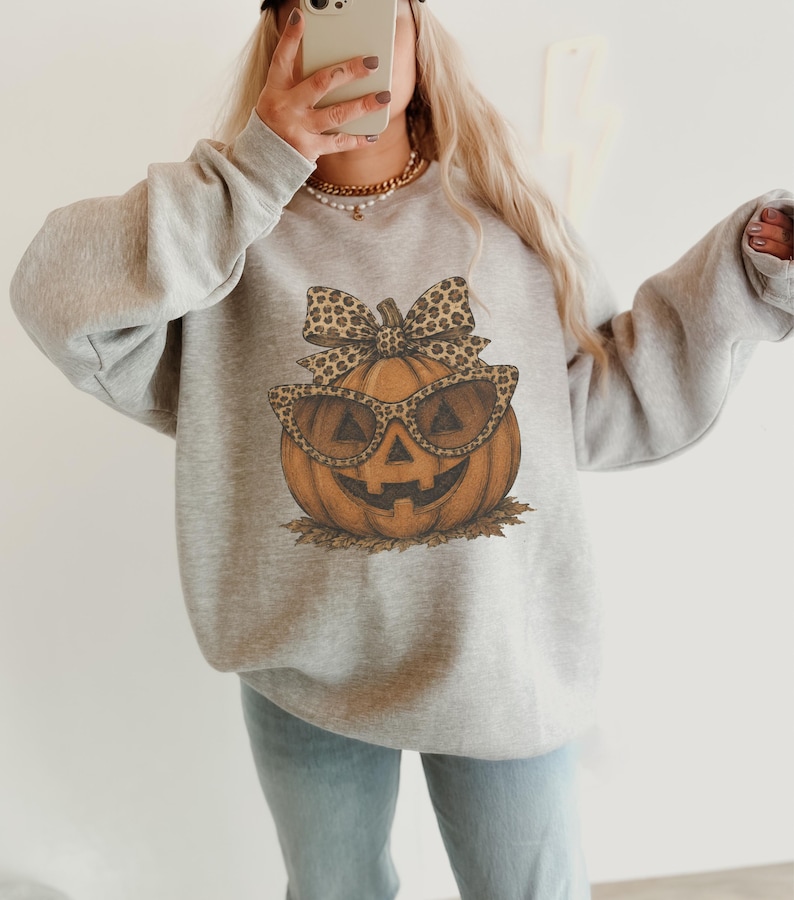Op de afbeelding: Een lichtgrijs sweatshirt met ronde hals en een centrale afbeelding van een oranje pompoen met een zonnebril met luipaardprint en een bijpassende strik. De pompoen rust op een klein hoopje bruine herfstbladeren.