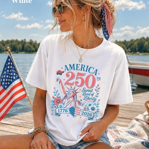 K&ouml;nnte beinhalten: Wei&szlig;es T-Shirt mit einem patriotischen Design zum 250. Jahrestag Amerikas. Die Grafik zeigt die Freiheitsstatue, florale Elemente und die Jahreszahlen 1776 und 2026. Das T-Shirt wird im Freien in der N&auml;he eines Sees mit einer amerikanischen Flagge getragen.
