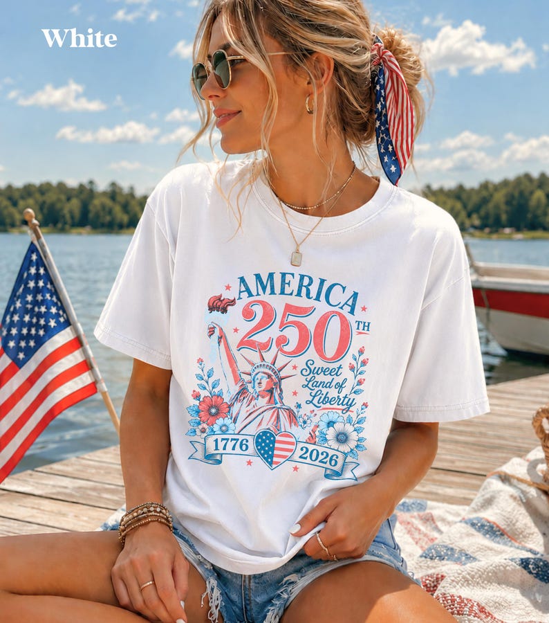 K&ouml;nnte beinhalten: Wei&szlig;es T-Shirt mit einem patriotischen Design zum 250. Jahrestag Amerikas. Die Grafik zeigt die Freiheitsstatue, florale Elemente und die Jahreszahlen 1776 und 2026. Das T-Shirt wird im Freien in der N&auml;he eines Sees mit einer amerikanischen Flagge getragen.