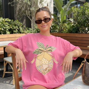 May include: A pink t-shirt with a lemon design featuring patchwork patterns, a green gingham leaf, and a yellow bow. The text "Crunchberry" is at the bottom. The shirt is worn by a person wearing sunglasses.