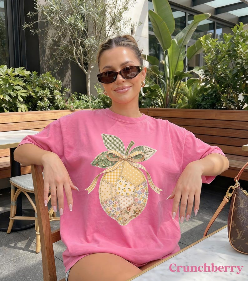 May include: A pink t-shirt with a lemon design featuring patchwork patterns, a green gingham leaf, and a yellow bow. The text "Crunchberry" is at the bottom. The shirt is worn by a person wearing sunglasses.
