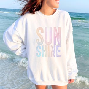 May include: White sweatshirt with the words "SUN SHINE" in pastel rainbow colors. The sweatshirt is long-sleeved and has a crew neck. The word "White" is printed at the bottom of the image.