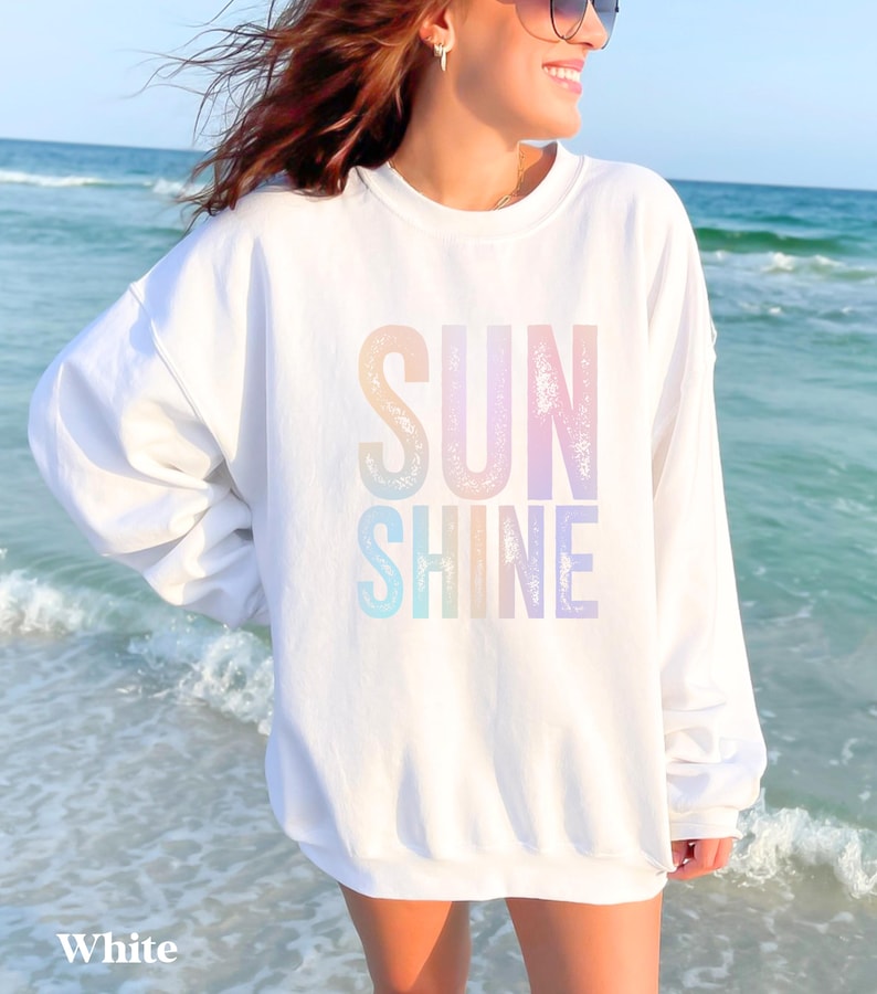 May include: White sweatshirt with the words "SUN SHINE" in pastel rainbow colors. The sweatshirt is long-sleeved and has a crew neck. The word "White" is printed at the bottom of the image.