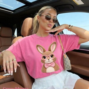 May include: A pink t-shirt featuring a crochet bunny wearing glasses and a bow tie. The word "Crunchberry" is printed at the bottom. The shirt is paired with a white skirt and accessories.