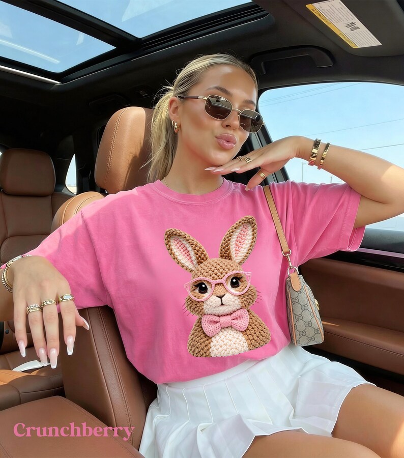 May include: A pink t-shirt featuring a crochet bunny wearing glasses and a bow tie. The word "Crunchberry" is printed at the bottom. The shirt is paired with a white skirt and accessories.