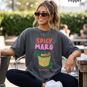 Puede incluir: Camiseta gris oscuro con las palabras "SPICY MARG" en rosa y naranja, y un gr&aacute;fico de una margarita con un chile. La camiseta tambi&eacute;n tiene las palabras "+EXTRA SPICY" debajo del gr&aacute;fico de la margarita.