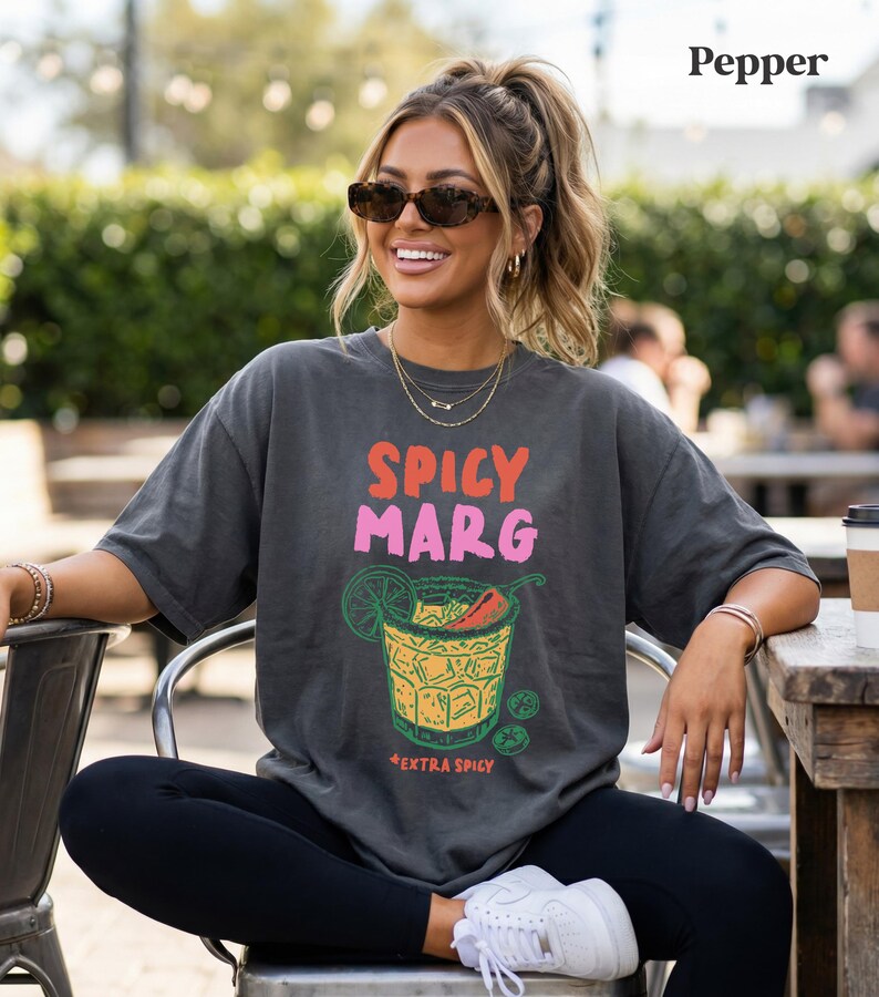 Puede incluir: Camiseta gris oscuro con las palabras "SPICY MARG" en rosa y naranja, y un gr&aacute;fico de una margarita con un chile. La camiseta tambi&eacute;n tiene las palabras "+EXTRA SPICY" debajo del gr&aacute;fico de la margarita.