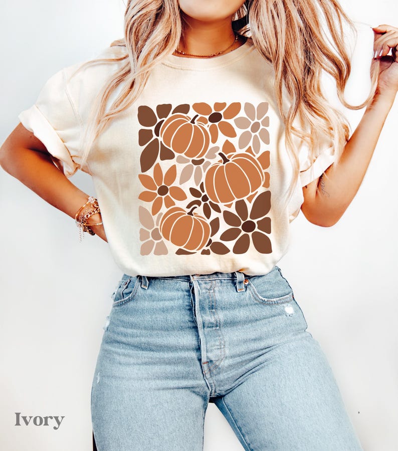 May include: Ivory-colored t-shirt featuring a fall-themed design. The graphic includes three pumpkins and floral elements in shades of brown and tan. The shirt is paired with light blue jeans. The word "Ivory" is printed at the bottom.