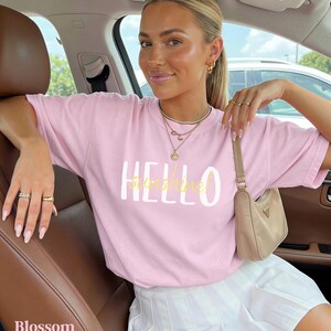 May include: A pale pink t-shirt with the text "HELLO SUNSHINE" in white and yellow. The outfit includes a white pleated skirt and a small beige handbag. The word "Blossom" is visible at the bottom left of the image.