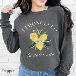 May include: A dark gray long-sleeve sweatshirt with a watercolor design of lemons and leaves. The text "LIMONCELLO" and "la dolce vita" are printed in gold. The word "Pepper" is printed in the bottom left corner.