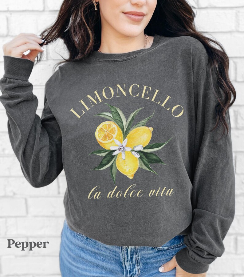 May include: A dark gray long-sleeve sweatshirt with a watercolor design of lemons and leaves. The text "LIMONCELLO" and "la dolce vita" are printed in gold. The word "Pepper" is printed in the bottom left corner.