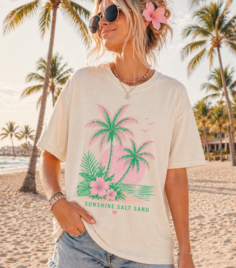 May include: Cream-coloured t-shirt with a tropical design of pink and green palm trees, a pink sun, and the text "SUNSHINE SALT SAND." The shirt has a relaxed fit and short sleeves, ideal for a beach holiday.