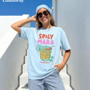 Puede incluir: Camiseta azul claro con la frase "Spicy Marg" en rojo y rosa, y un gr&aacute;fico de c&oacute;ctel con lima y chile. La camiseta tambi&eacute;n tiene las palabras "Extra Spicy" debajo del c&oacute;ctel.