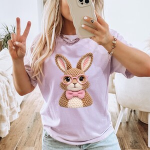 May include: A light purple t-shirt featuring a detailed illustration of a brown bunny wearing pink glasses and a pink bow tie. The bunny has pink-lined ears. The word "Orchid" is printed in the upper right corner.