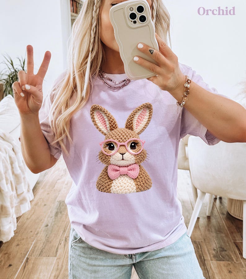 May include: A light purple t-shirt featuring a detailed illustration of a brown bunny wearing pink glasses and a pink bow tie. The bunny has pink-lined ears. The word "Orchid" is printed in the upper right corner.