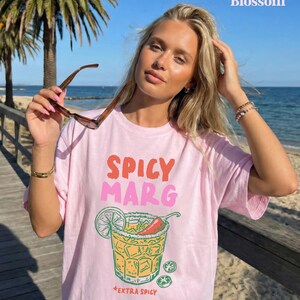 Puede incluir: Camiseta rosa claro con las palabras "SPICY MARG" en naranja y rosa. Debajo del texto hay un gr&aacute;fico de una margarita con lima, chile y sal. Las palabras "EXTRA SPICY" est&aacute;n debajo del gr&aacute;fico.