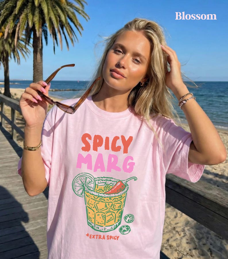 Puede incluir: Camiseta rosa claro con las palabras "SPICY MARG" en naranja y rosa. Debajo del texto hay un gr&aacute;fico de una margarita con lima, chile y sal. Las palabras "EXTRA SPICY" est&aacute;n debajo del gr&aacute;fico.