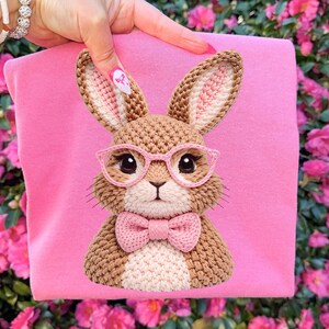 May include: A pink fabric with a crocheted bunny design. The bunny has brown fur, pink glasses, and a pink bow tie. The bunny's ears have a pink interior. The background is a vibrant display of pink flowers.