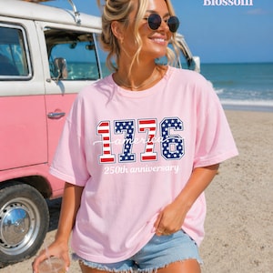 May include: A pale pink t-shirt featuring the text "1776 America 250th anniversary" in red, white, and blue. The shirt is worn by a person on a beach, with a vintage pink and white van in the background.