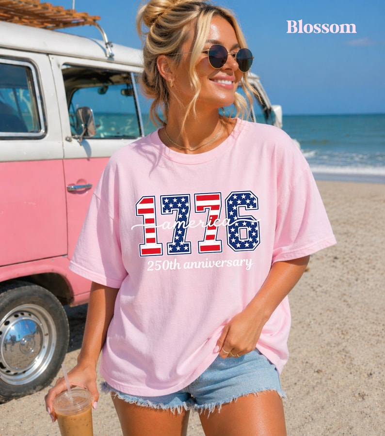 May include: A pale pink t-shirt featuring the text "1776 America 250th anniversary" in red, white, and blue. The shirt is worn by a person on a beach, with a vintage pink and white van in the background.