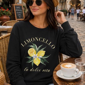 May include: Black long-sleeve sweatshirt with the word "Limoncello" in gold script above a watercolor illustration of lemons and leaves. The phrase "la dolce vita" is printed below the illustration. A cup of coffee sits on a wooden table.