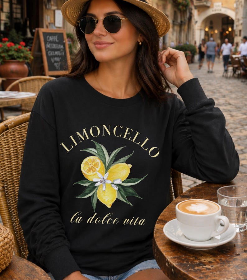 May include: Black long-sleeve sweatshirt with the word "Limoncello" in gold script above a watercolor illustration of lemons and leaves. The phrase "la dolce vita" is printed below the illustration. A cup of coffee sits on a wooden table.