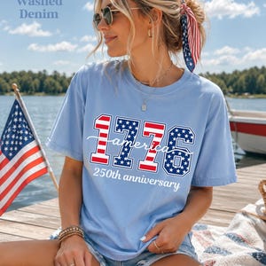 May include: Pale blue t-shirt featuring the text "1776 America 250th anniversary" in red, white, and blue. The shirt is worn by a person with sunglasses and a patriotic hair accessory. An American flag is in the background.