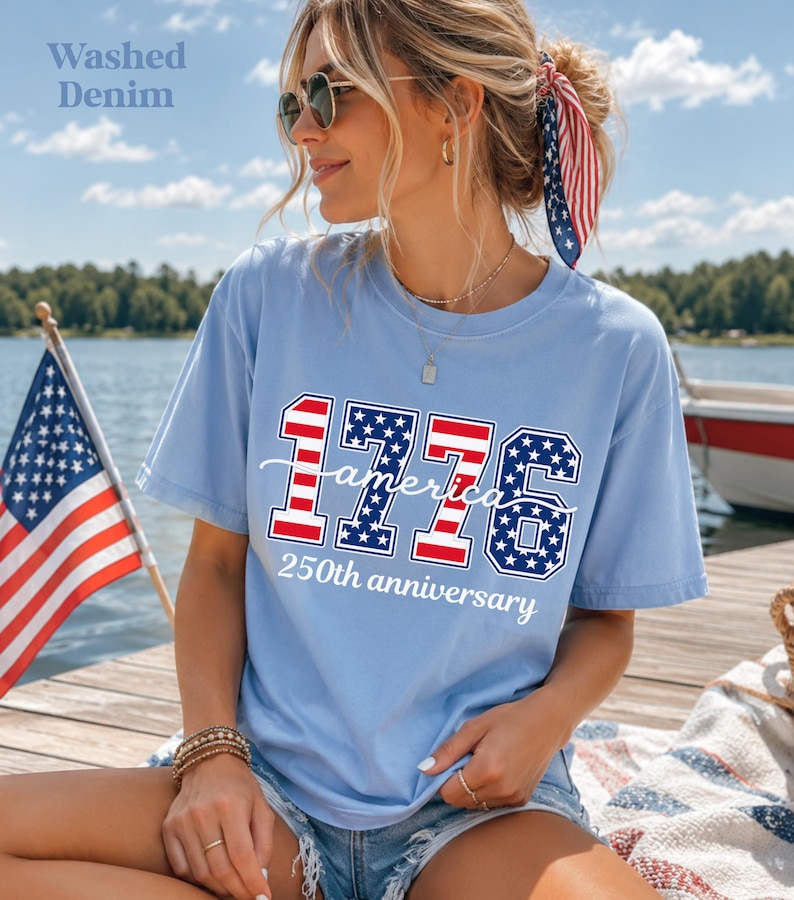 May include: Pale blue t-shirt featuring the text "1776 America 250th anniversary" in red, white, and blue. The shirt is worn by a person with sunglasses and a patriotic hair accessory. An American flag is in the background.