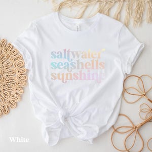 May include: White t-shirt with the words "saltwater seashells sunshine" in pastel colors. The shirt is knotted at the waist. The word "White" is at the bottom left of the shirt.