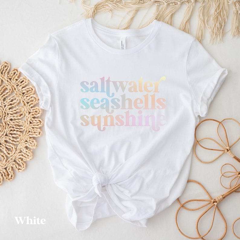 May include: White t-shirt with the words "saltwater seashells sunshine" in pastel colors. The shirt is knotted at the waist. The word "White" is at the bottom left of the shirt.