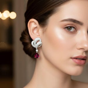 May include: A close-up of a silver earring with a swirling design, set with clear crystals. Each earring has a teardrop-shaped, deep red gemstone pendant. The earrings are designed to catch the light and sparkle.