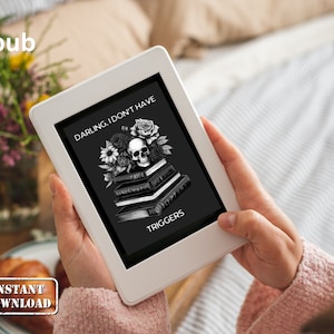 Puede incluir: Una lectora electrónica blanca que muestra la portada de un libro titulado "Darling, I Don't Have Triggers". La portada presenta una ilustración en blanco y negro de una calavera con flores y una pila de libros.