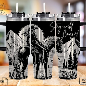 May include: A stainless steel tumbler with a black and white graphic of a cowgirl riding a horse. The tumbler has a black lid and a straw. The text "Stay Wild Cowgirl" is printed on the tumbler.