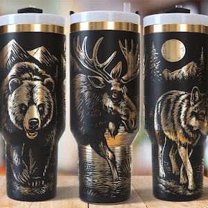 May include: Three black travel mugs with gold animal illustrations: a bear, a moose, and a wolf. Each mug has a black handle, a white lid, and a gold rim. The mugs are decorated with mountain and forest scenes.