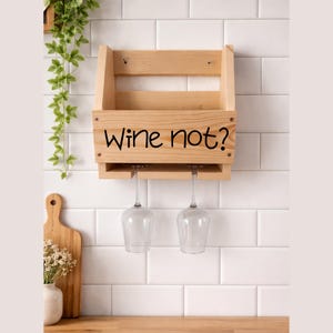 Puede incluir: Un botellero de madera clara con la frase "Wine not?" en letras negras. Dos copas de vino cuelgan debajo. El botellero est&aacute; montado en una pared de azulejos blancos. Una tabla de cortar de madera y un jarr&oacute;n con flores est&aacute;n en primer plano.